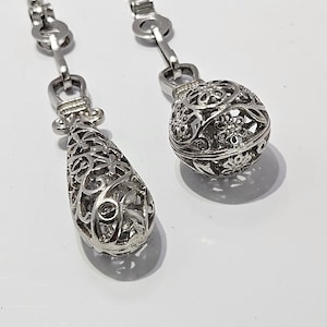 May include: Two silver filigree pendant charms, one shaped like a teardrop and the other a sphere, hanging from silver chains.