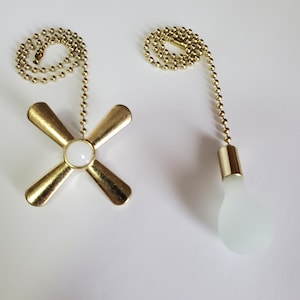 Puede incluir: Dos cadenas de tirador de luz doradas. Una cadena tiene un tirador de aspa de ventilador de plástico blanco. La otra cadena tiene un tirador de bombilla de plástico blanco.