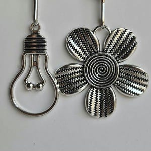 May include: Silver-toned earrings. One earring is a lightbulb shape, the other is a flower with a spiral center and zigzag petal detailing. Both earrings hang from a simple hook.