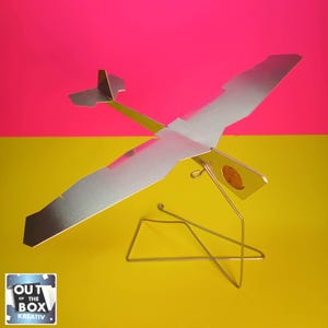 Pode incluir: Um modelo de avião prateado com detalhes em amarelo e verde é exibido em um suporte de arame. O avião tem uma envergadura de aproximadamente 30 cm. O fundo é dividido em seções rosa brilhante e amarelo, com o texto "OUT OF THE BOX KREATIV" no canto inferior esquerdo.
