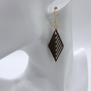 May include: A single earring with a geometric design is displayed. The earring is a dark brown diamond shape with a series of angled lines cut into the wood. The earring hangs from a gold-colored hook. The background is white.