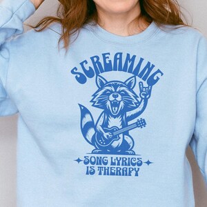 Può includere: Felpa blu chiaro con un'immagine di un procione che suona la chitarra e il testo "Screaming Song Lyrics Is Therapy".
