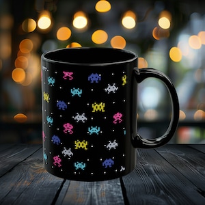 May include: Black ceramic coffee mug with a pattern of colorful pixelated space invaders. The mug has a white handle.