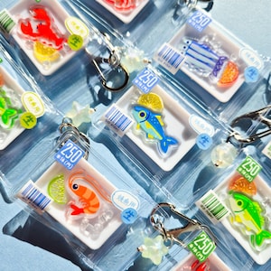 Könnte beinhalten: Eine Sammlung von durchsichtigen Kunststoff-Schlüsselanhängern mit Miniatur-Lebensmittelschalen und Acryl-Meeresfrüchte-Anhängern, darunter Hummer, Garnelen und verschiedene Fische, kombiniert mit Zitrusfrüchten, Barcodes und Preisschildern mit der Aufschrift 250.