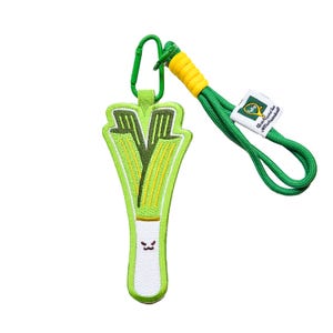 May include: A green and white leek keyring with a smiling face. The keyring has a green lanyard with a yellow and green tag that says "Black Forest Ham & Sausage Kitchen".