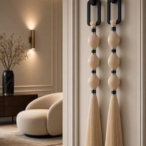 Può includere: Due decorazioni da parete verticali con ganci rettangolari neri nella parte superiore. Ogni pezzo è composto da tre ornamenti sferici in fibra color crema impilati, separati da sottili fasce nere e terminanti con lunghe nappe fluide.