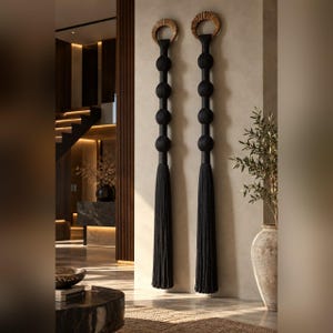 May include: Two long, vertical wall hangings featuring a series of four black spherical fabric-covered beads stacked below a circular wooden ring, finished with a long, thick black tassel at the bottom, suspended against a neutral wall.
