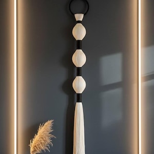 Puede incluir: Un colgante de pared decorativo con un anillo negro, tres elementos blancos ovalados tejidos y una larga borla blanca. Los elementos están conectados por secciones cilíndricas negras. La pared es de color gris oscuro y se ve un jarrón con plantas secas.