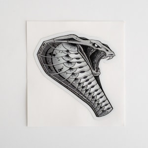 Puede incluir: Una ilustración en blanco y negro de una cobra con la boca abierta, mostrando sus colmillos. Las escamas de la serpiente son detalladas y texturizadas.