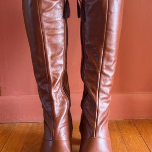 May include: A pair of knee-high brown leather boots. The boots have a pointed toe, a zipper with a tassel, and a slight heel. The leather has a smooth, shiny finish. The boots are standing on a wooden floor.