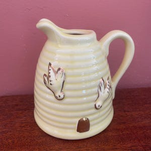 May include: A small, yellow ceramic creamer in the shape of a beehive with two gold birds on the side. The creamer has a gold rim and a gold handle.