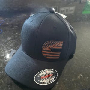 May include: Navy blue Flexfit baseball cap with the Cummins logo in brown and orange, featuring a star design. The cap has a curved peak and a Flexfit sticker. The cap is size L/XL.