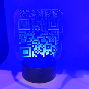 Può includere: Una luce blu con un pannello in acrilico trasparente che dice "SCAN ME" e ha un codice QR. La luce è su una base nera.