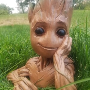 May include: A brown and green ceramic planter shaped like a character from the movie Guardians of the Galaxy. The planter is sitting in a bed of green grass.