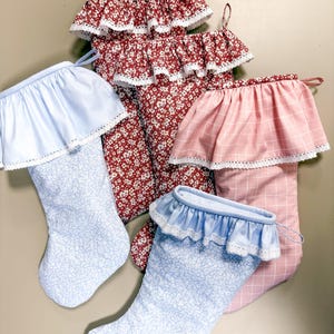 Puede incluir: Una colección de calcetines navideños en varios patrones y colores. Los calcetines tienen una parte superior con volantes y ribete de encaje. Los patrones incluyen un estampado floral rojo, un estampado floral azul claro y un patrón de cuadros rosas.