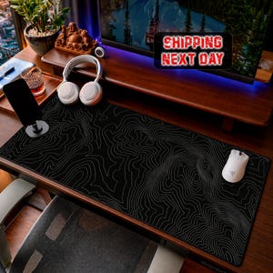Peut inclure: Un tapis de bureau rectangulaire noir orné d'un motif de lignes topographiques blanches. Le tapis est posé sur un bureau en bois sombre avec une souris d'ordinateur sans fil blanche, un casque audio blanc, un support de smartphone et un verre.