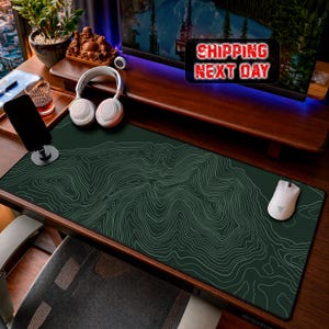 May include: A rectangular desk mat featuring a dark green surface with a white topographic map contour line pattern. The mat is positioned on a wooden desk surface alongside a white computer mouse, white over-ear headphones, and a black smartphone stand.