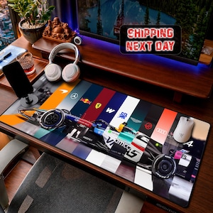 May include: A rectangular desk mat featuring a Formula 1 race car graphic overlaid on a background of vertical color stripes representing various racing team logos, including McLaren, Mercedes, Red Bull, Ferrari, Williams, and Audi.