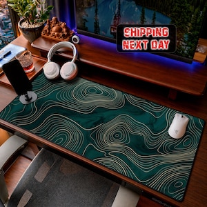 May include: A rectangular desk mat featuring a dark green marble-like background with thin, gold-coloured topographic contour lines. The mat is positioned on a wooden desk surface alongside a white computer mouse, white headphones, and a black phone stand.