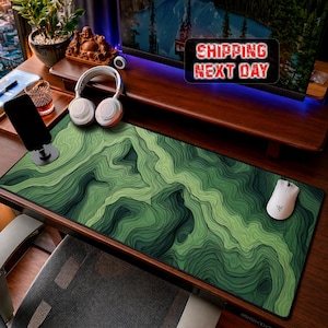 Può includere: Un tappetino da scrivania rettangolare con un motivo a mappa topografica verde e nero con linee di contorno stratificate. Il tappetino è posizionato su una scrivania in legno accanto a un mouse bianco, cuffie bianche e un supporto per smartphone.
