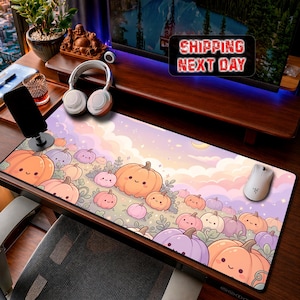 Op de afbeelding: Een rechthoekige bureaumat met een grillige illustratie van talloze lachende pompoenen in tinten oranje, roze en paars tegen een zachte pastelkleurige lucht met wolken. De mat ligt op een houten bureau met een witte computermuis.