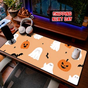 May include: A rectangular desk mat featuring a light orange background with a repeating pattern of white ghosts, orange pumpkins with black facial features, black bats, and small black stars. A white computer mouse rests on the right side.