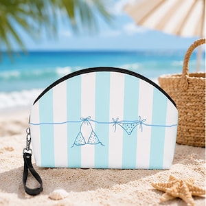 May include: A semi-circular zippered pouch featuring a light blue and white vertical striped pattern. The front displays a simple line drawing of a two-piece bikini hanging on a clothesline. A black wrist strap is attached to the side zipper.