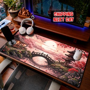 May include: A rectangular desk mat featuring a printed illustration of a Japanese landscape with a pagoda, a stone bridge over a river, cherry blossom trees, and a large sun in the background. The colour palette consists of soft pinks, oranges, and dark tones.
