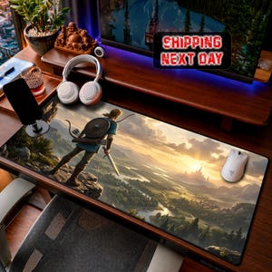 May include: A rectangular desk mat featuring a printed landscape illustration of a figure standing on a cliff edge overlooking a valley with a distant castle at sunset. The mat is positioned on a wooden desk surface with a white computer mouse on top.