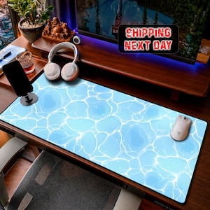 May include: A rectangular desk mat featuring a light blue water surface pattern with white caustic light reflections. The mat is positioned on a wooden desk surface alongside a white computer mouse, a pair of white headphones, and a black smartphone stand.