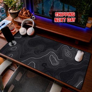 Peut inclure: Un tapis de bureau rectangulaire présentant une surface noire avec des lignes de contour topographiques blanches et grises. Le tapis est placé sur un bureau en bois avec une souris d'ordinateur blanche, un casque audio blanc et un support de smartphone noir.