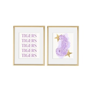May include: Two framed prints with a gold frame. The print on the left has the word "TIGERS" repeated five times in a light purple color. The print on the right has a purple cheetah with gold stars on a light purple background.