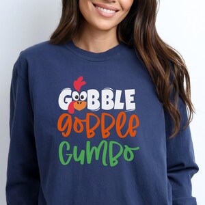 May include: A navy blue long-sleeved shirt with a white, orange, and green graphic that says "Gobble gobble gumbo" with a cartoon turkey head.