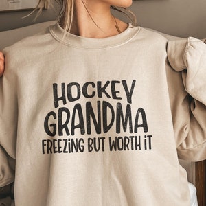 May include: A beige sweatshirt with black text that reads 'Hockey Grandma Freezing But Worth It.'