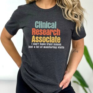 May include: A dark gray t-shirt with the text "Clinical Research Associate I don't have trust issues just a lot of monitoring visits" in a retro font with colorful accents.