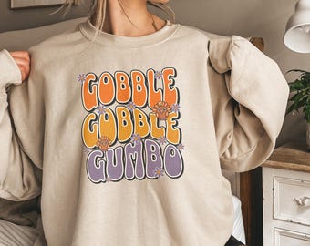 May include: Beige sweatshirt featuring the words "GOBBLE GOBBLE GUMBO" in orange, yellow, and purple. The text has a retro design with floral accents and turkey illustrations. A comfortable, casual top.