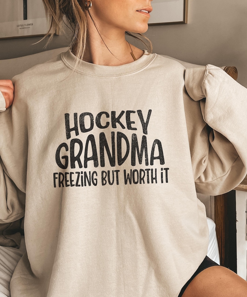 May include: A beige sweatshirt with black text that reads 'Hockey Grandma Freezing But Worth It.'