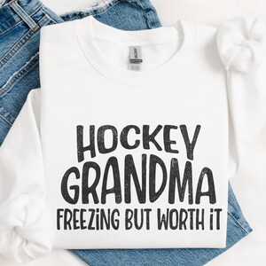 May include: White sweatshirt with black text that reads "Hockey Grandma Freezing But Worth It".
