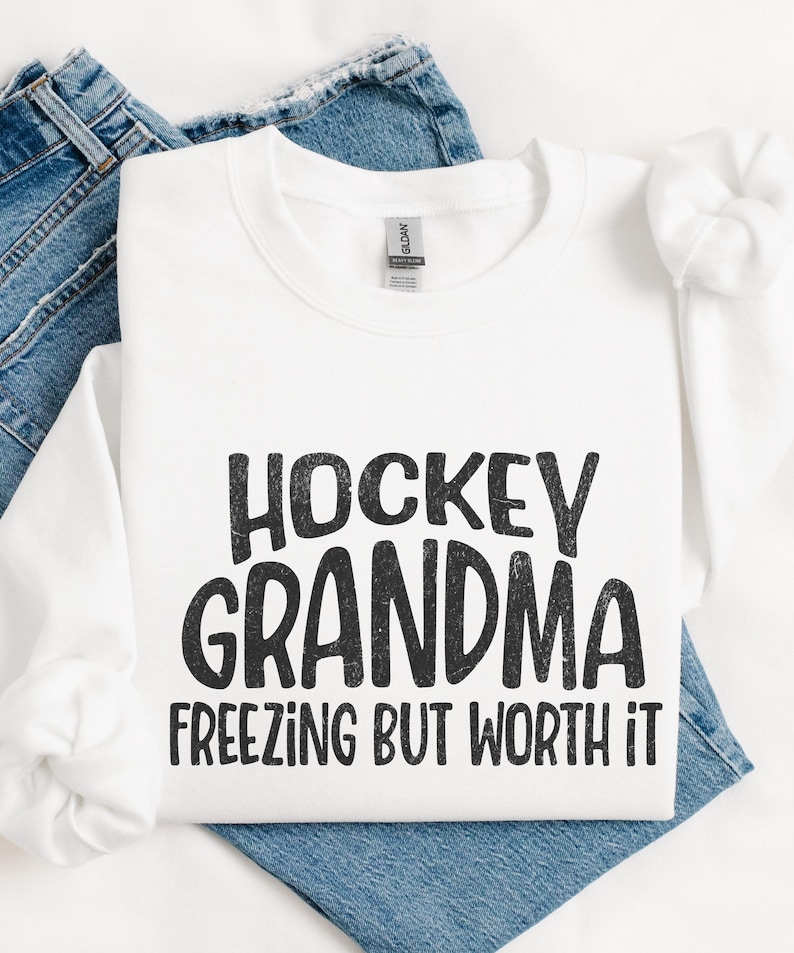 May include: White sweatshirt with black text that reads "Hockey Grandma Freezing But Worth It".