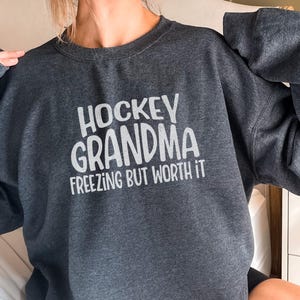 May include: A dark gray sweatshirt with the text "Hockey Grandma Freezing But Worth It" in white.