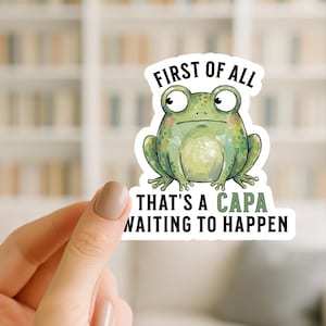 May include: A sticker featuring a cartoon frog with the text "FIRST OF ALL THAT'S A CAPA WAITING TO HAPPEN". The frog is green with a light green belly and pink cheeks. The text is in black and green.