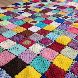 Könnte beinhalten: Eine große, handgefertigte Häkeldecke, die aus zahlreichen bunten Quadraten besteht. Die Quadrate sind in verschiedenen Farbtönen von Rot, Blau, Grün, Gelb, Lila und Braun gehalten. Die Decke hat ein Patchwork-Design und wirkt lebendig und strukturiert.