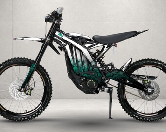 May include: An electric dirt bike featuring a white and black frame with a teal gradient graphic pattern. The bike includes a black seat, black knobby tires, front and rear disc brakes, a kickstand, and a suspension system with a central shock.