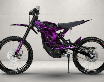 May include: An electric dirt bike featuring a black frame with vibrant purple graphic decals. The bike includes a black seat, knobby tires, front and rear disc brakes, a kickstand, and a suspension system. The frame displays a stylized monster graphic.