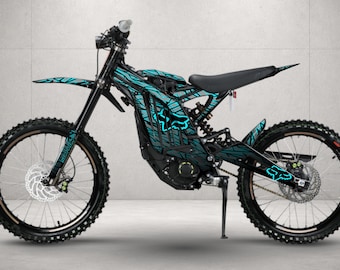 May include: An electric dirt bike featuring a black frame with a cyan blue graphic wrap. The design includes a repeating geometric pattern and the Fox Racing logo in cyan. It has black spoked wheels, disc brakes, a black seat, and a kickstand.