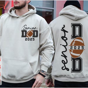 May include: A beige hoodie with the text "Senior Dad 2025" and a football graphic on the front. The back of the hoodie has the text "Senior Dad" with a football graphic and the year "2025" on the football.
