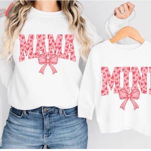 May include: Two white shirts with pink heart print designs. The larger shirt says "MAMA" with a pink bow. The smaller shirt says "MINI" with a pink bow.