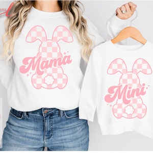 May include: Two white shirts with pink checkered bunny designs. The larger shirt says "Mama" and the smaller shirt says "Mini".