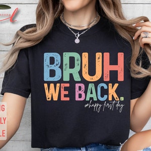 May include: A black t-shirt with the text "BRUH WE BACK." in rainbow colors with a distressed effect. The text "#happy first day" is written in smaller letters below the main text.