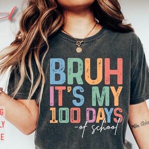 May include: A dark grey t-shirt with the text "BRUH IT'S MY 100 DAYS - of school" in colourful, distressed lettering.